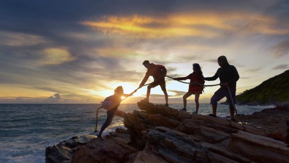 Male and female hikers climbing up silhouette mountain cliff and one of them giving helping hand. People helping and, team work concept.